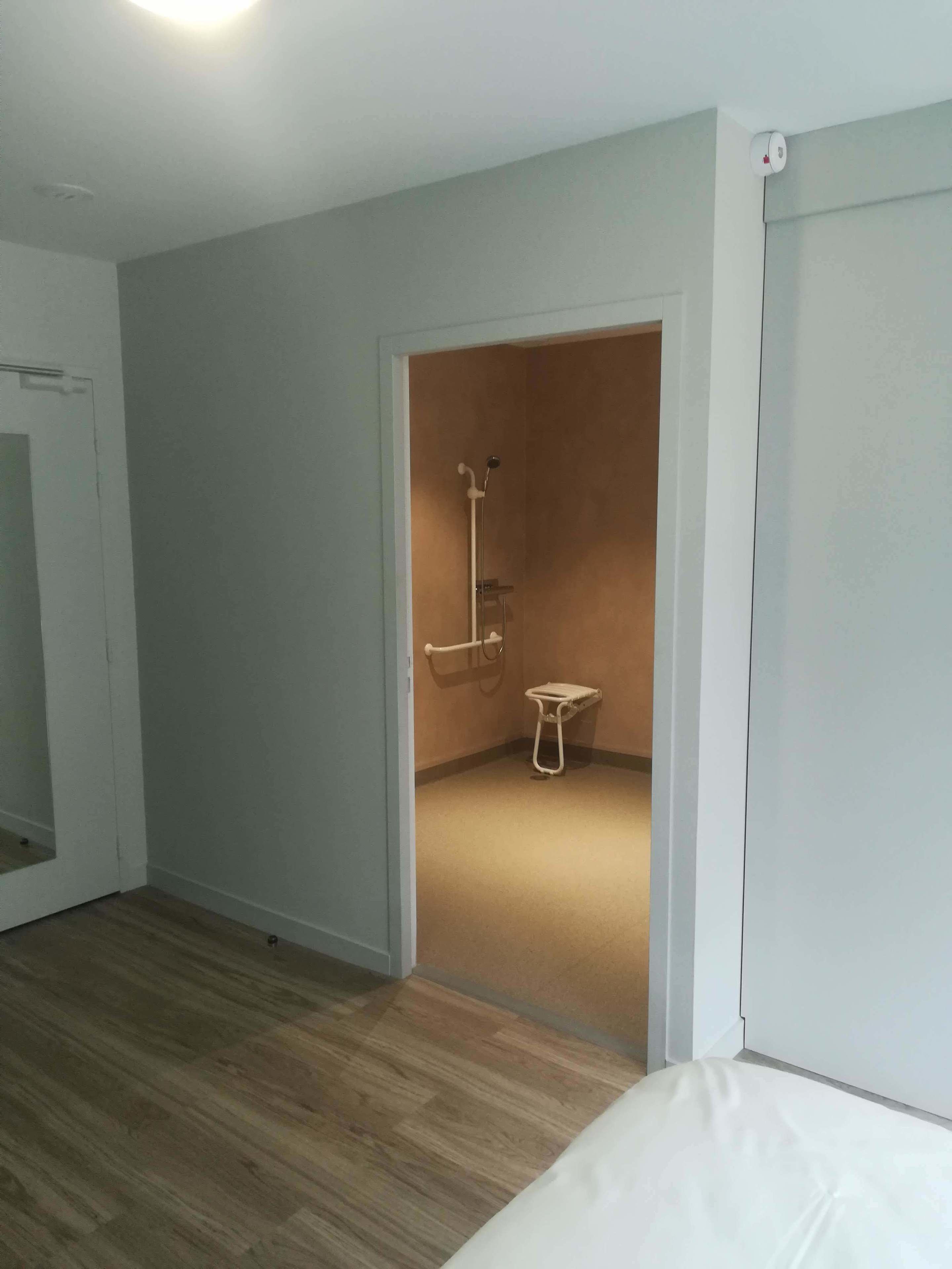 Une pièce avec un plancher en bois, un lit et un espace douche équipé d’un porte-serviettes et d’un petit tabouret.
