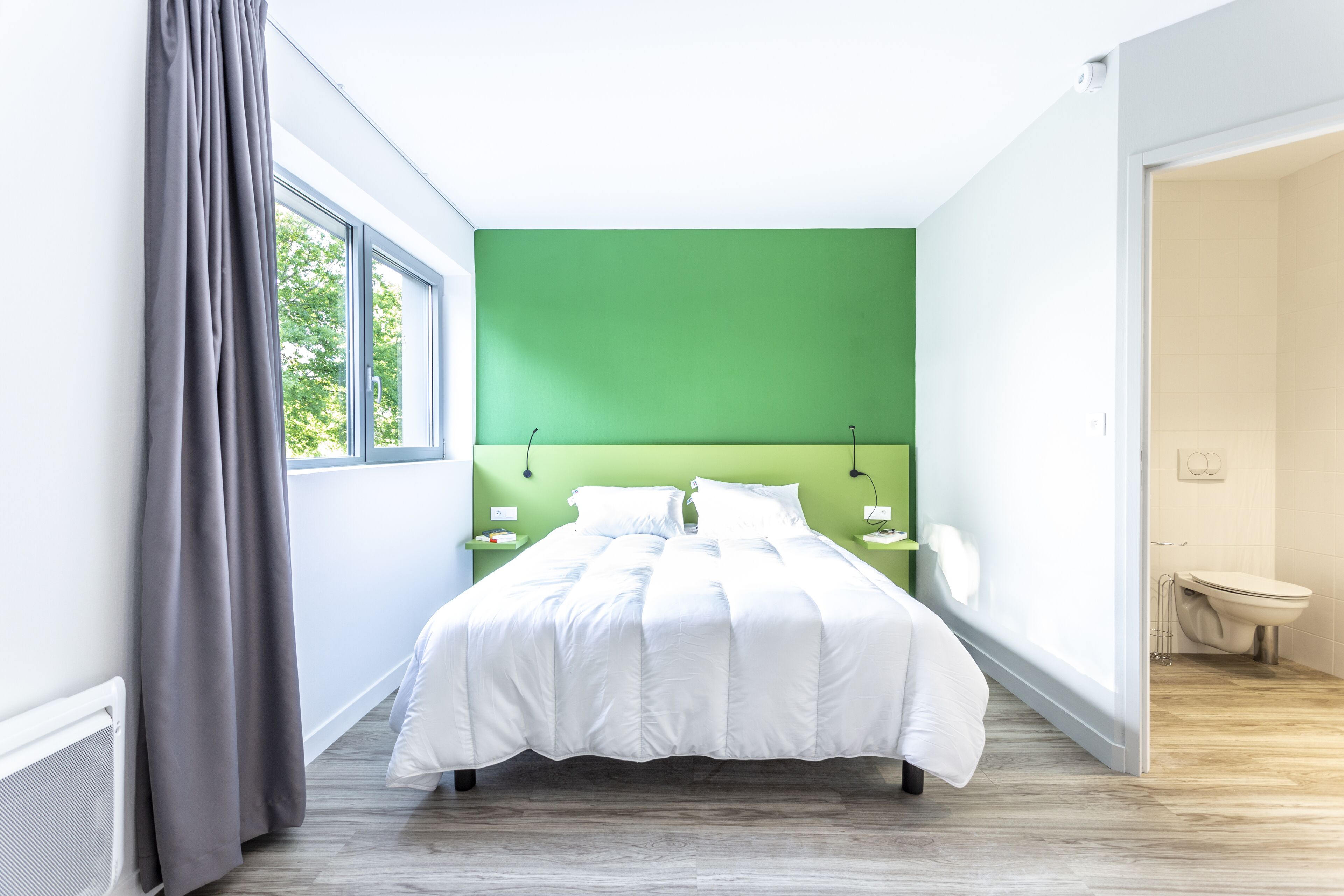 A modern bedroom with a green accent wall, a large bed, bedside tables, and a view of the outdoors.