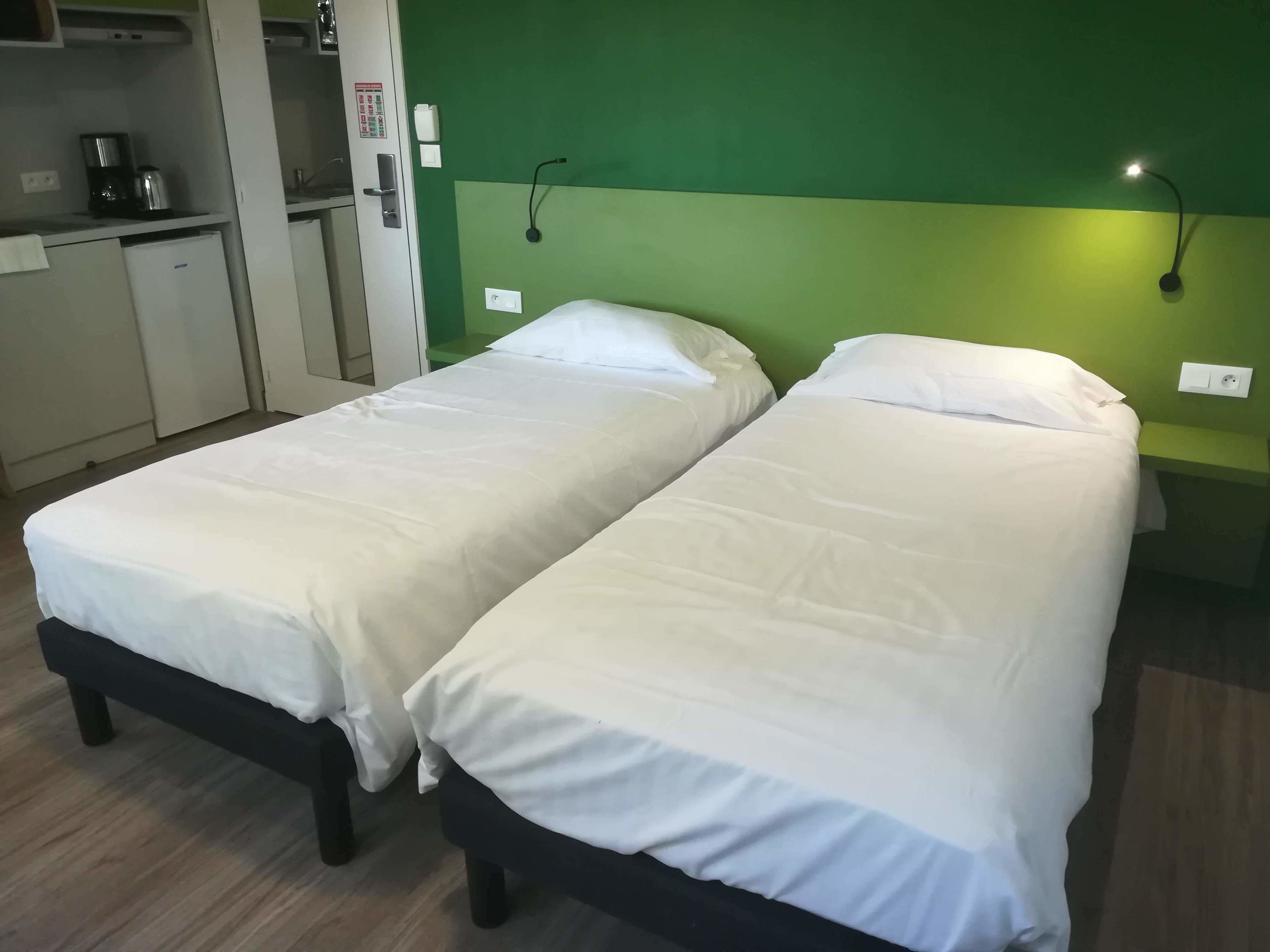 Quarto com duas camas de solteiro, cozinha compacta e parede de destaque verde.