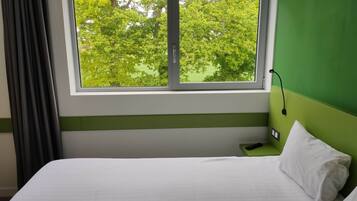 A hotel room with a bed, a window with a view of trees, a green wall, and a bedside table with a remote control.