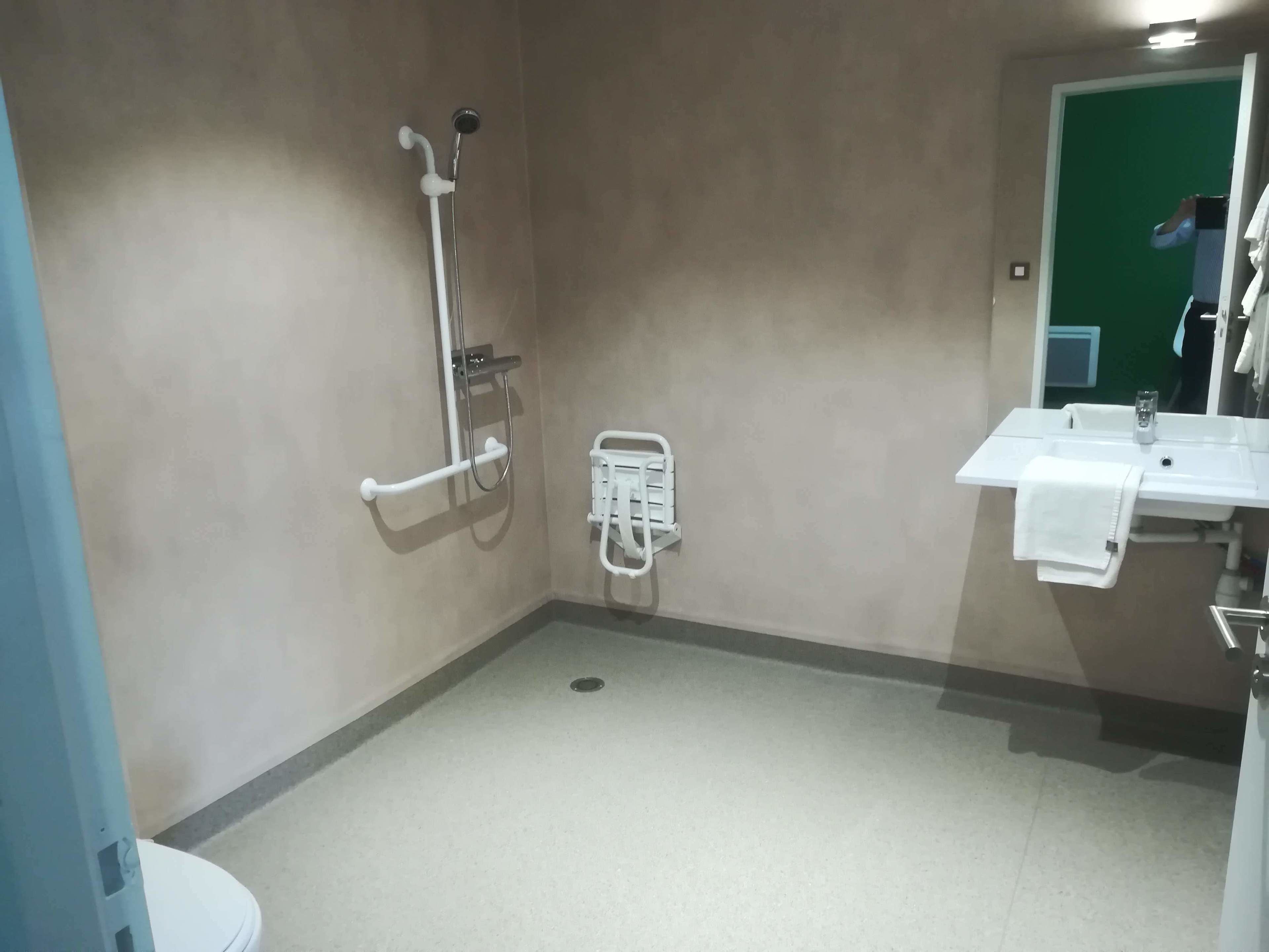A small, plain bathroom with a toilet, a shower with a grab bar, and a sink with a towel hanging on the wall.