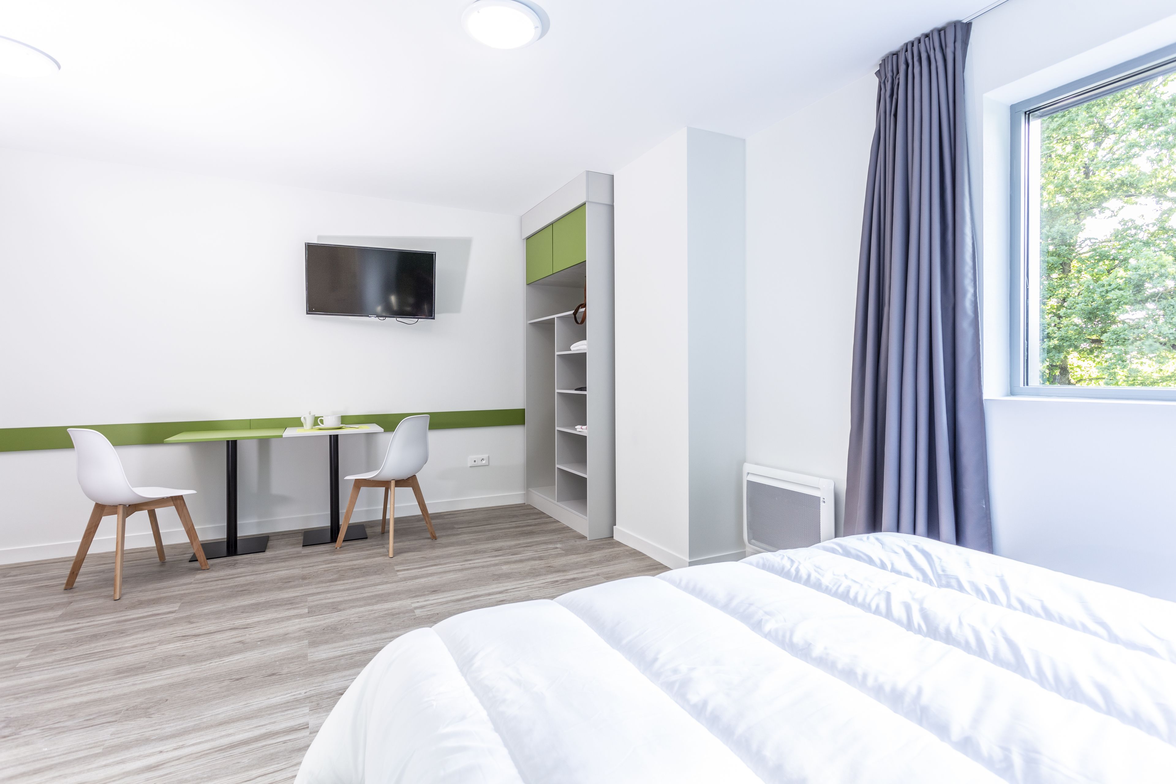 A hotel room with a bed, a desk with chairs, a TV mounted on the wall, and a window with curtains.