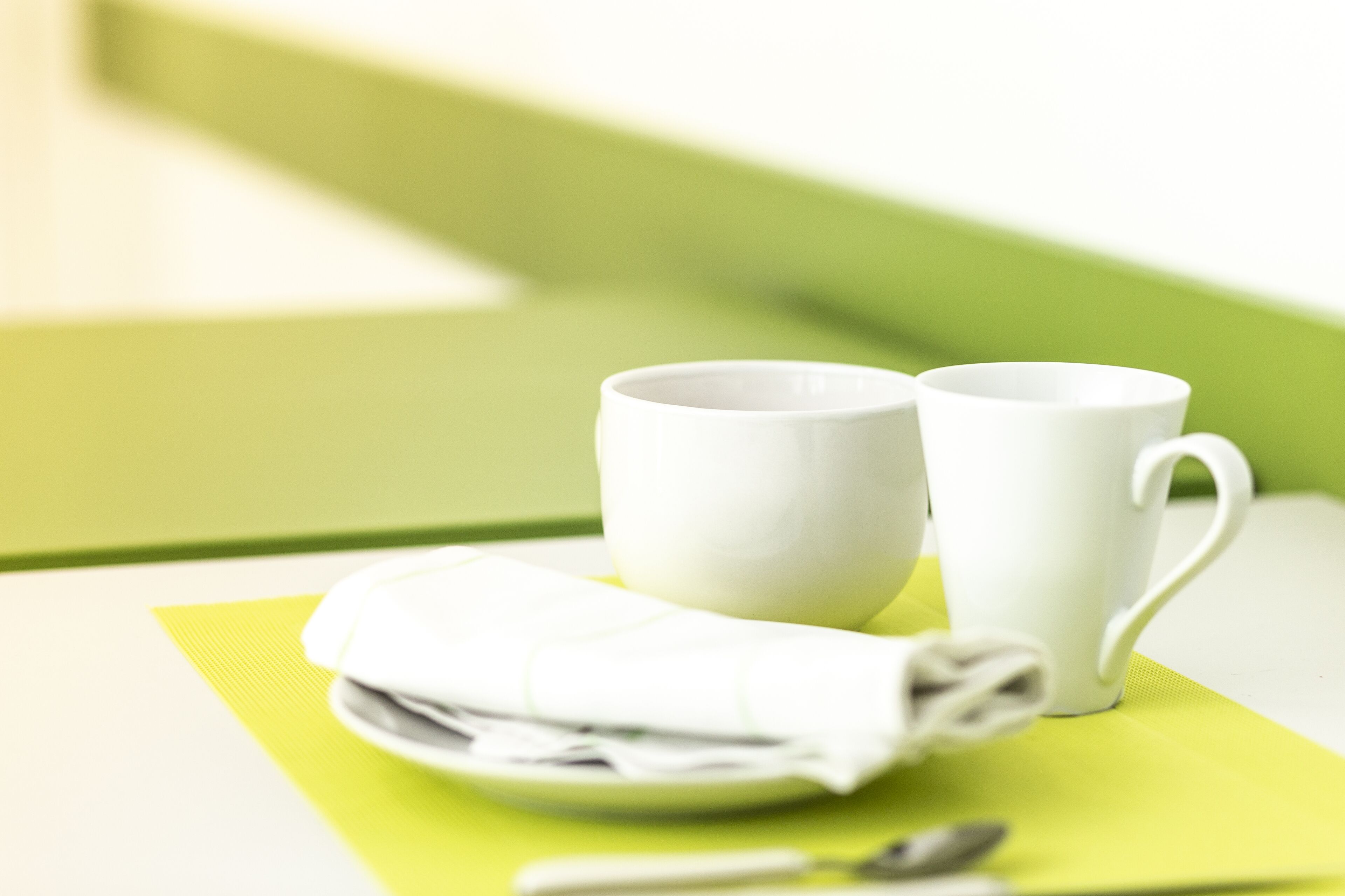 Una mesa con dos tazas blancas, una servilleta doblada y una cuchara sobre un mantel individual verde.