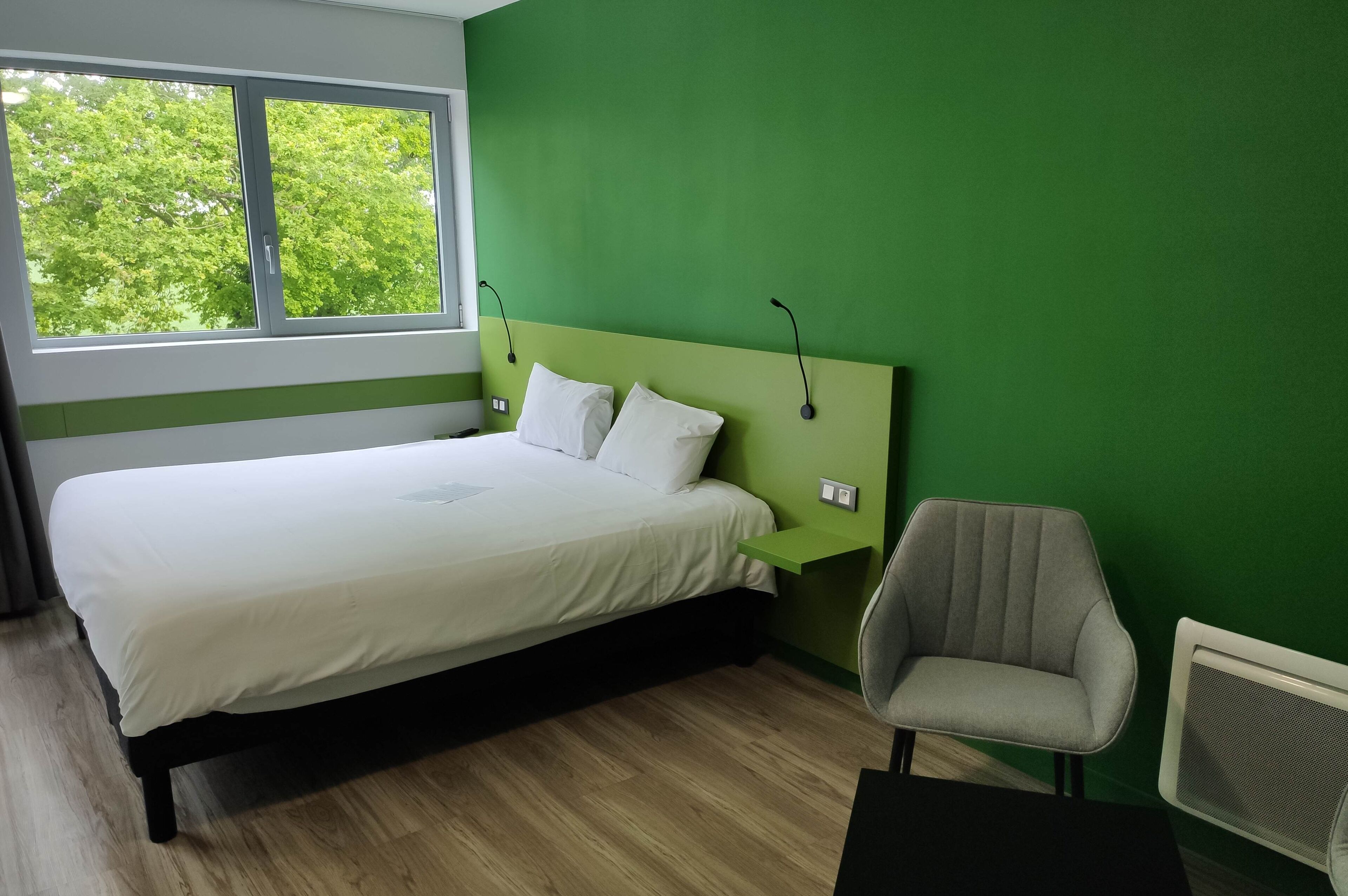 Ein Schlafzimmer mit grüner Wand, einem Bett mit weißer Bettwäsche, einem Sessel und einem Fenster mit Blick auf Bäume.