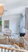 Um quarto com teto branco, uma luminária de fibra natural grande, uma cadeira de vime, uma mesa redonda, um pequeno banquinho, um espelho, uma porta e uma janela com cortinas.