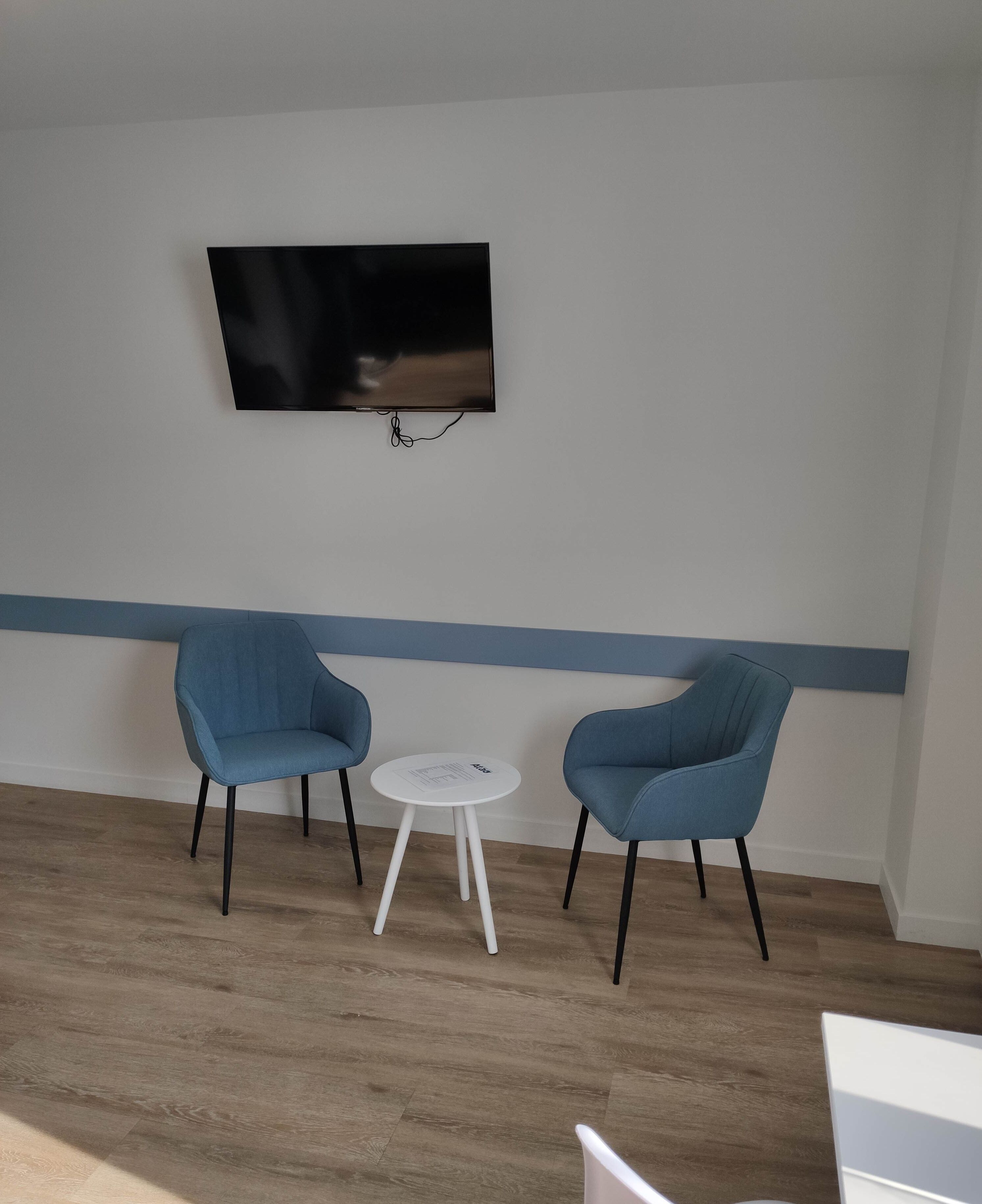 A room with a flat-screen TV mounted on the wall, two blue chairs, and a small white table.
