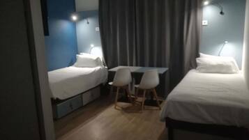 A small, well-lit room with two single beds, a desk, and two chairs.