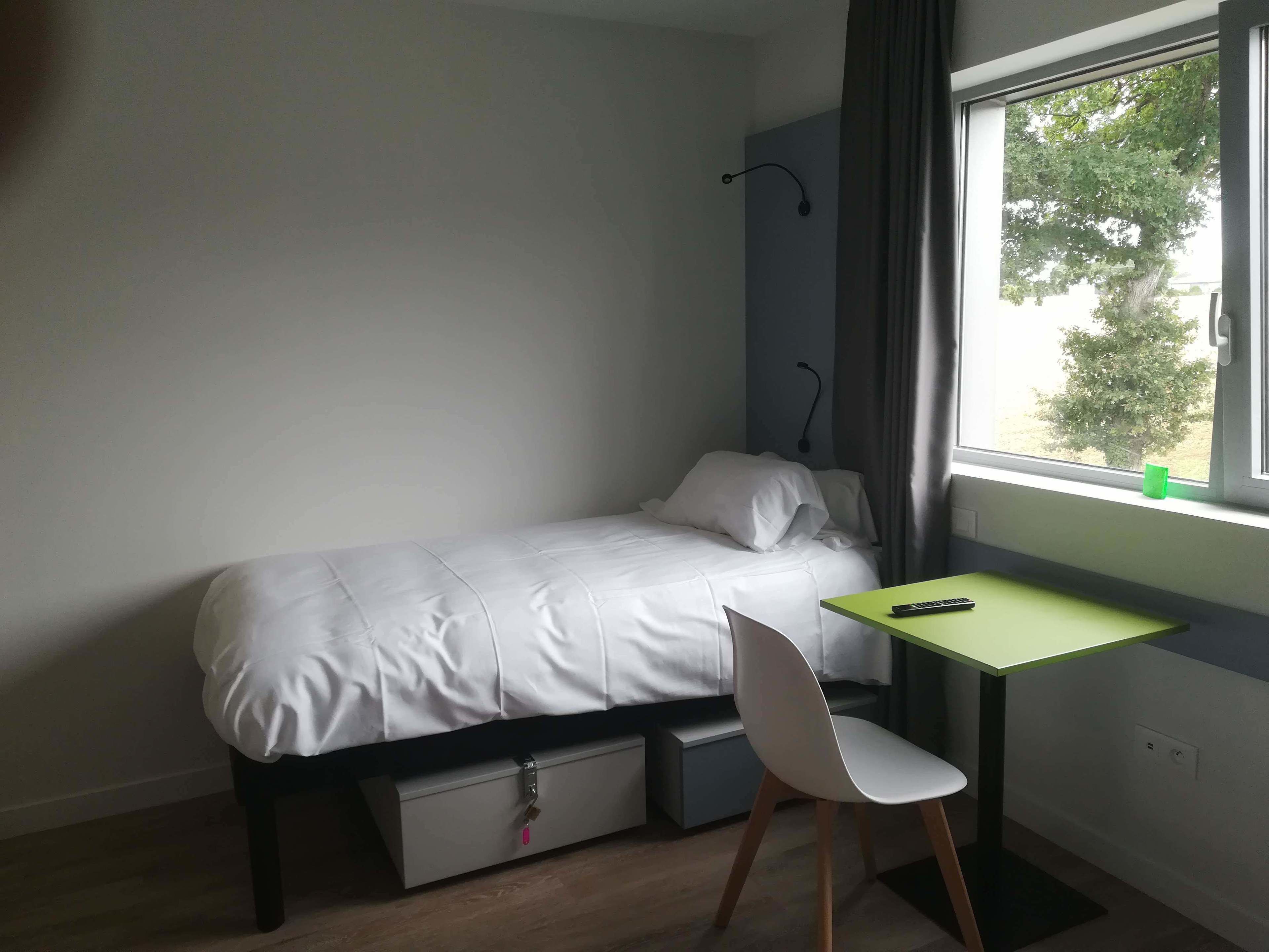 Een klein, netjes opgemaakt bed met een witte matras en kussens, een groen bureau met een stoel, en een raam met uitzicht op bomen.