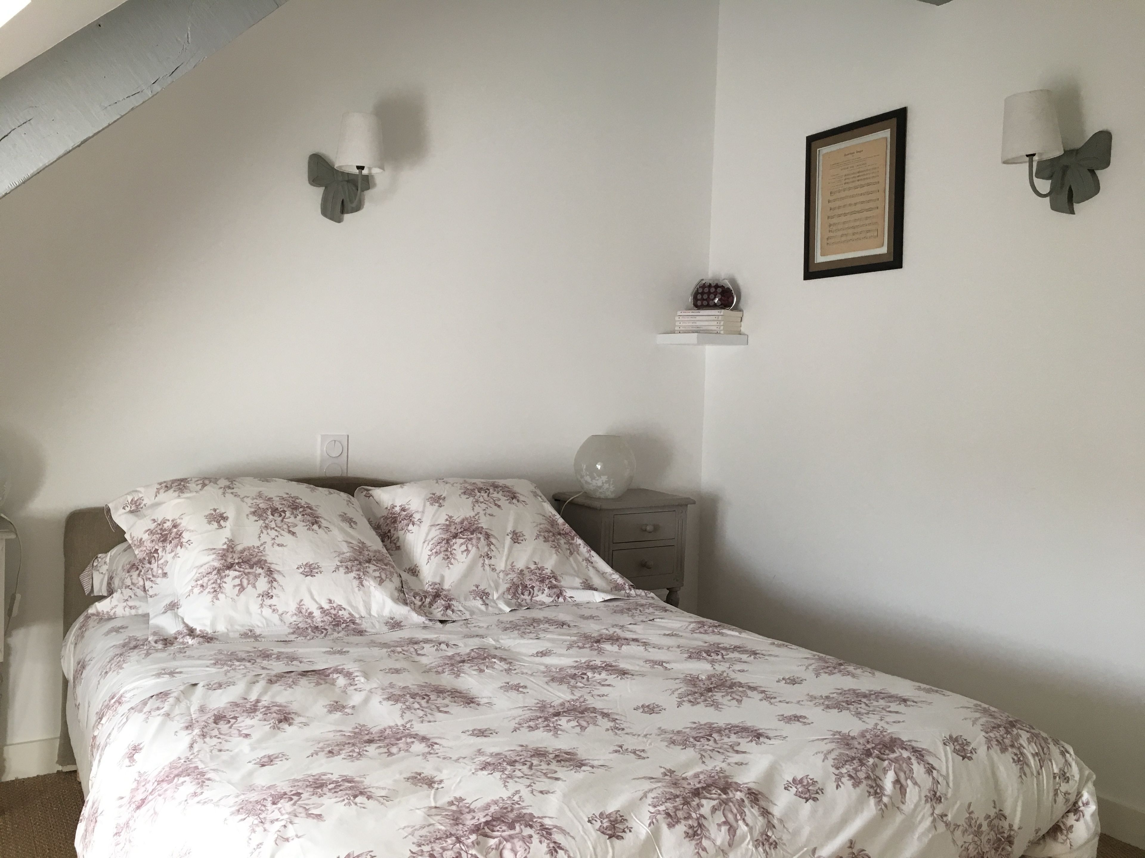 Een slaapkamer met een bed, een nachtkastje, een wandplank met boeken en twee wandlampen.