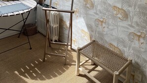 Una habitación con techo inclinado, un espejo redondo, una silla de mimbre, un radiador, una mesa con mantel de encaje y una ventana con persiana de madera.