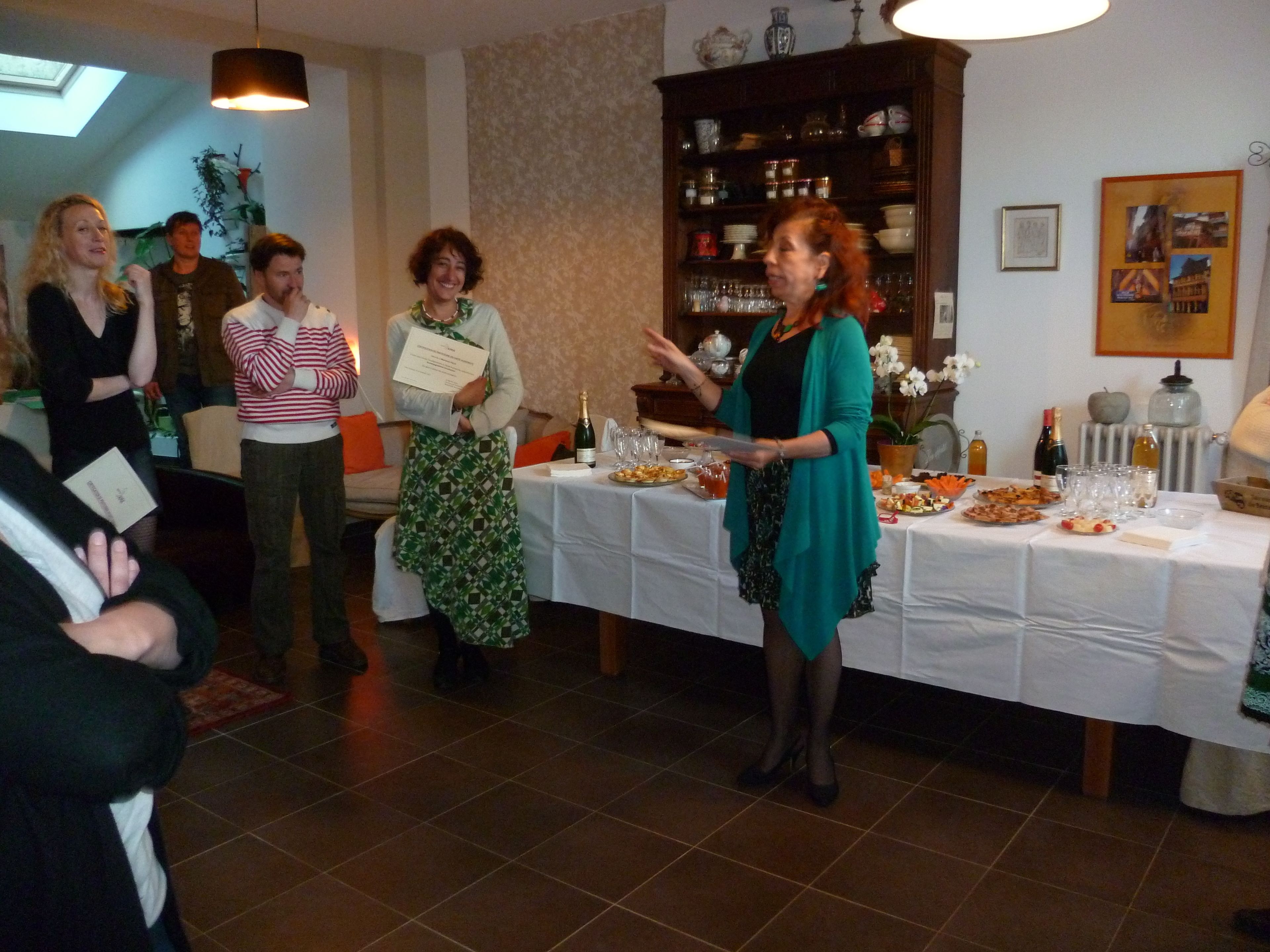 A group of people at a social event in a room with a tiled floor, a white table with food and drinks, and a bookshelf in the background.