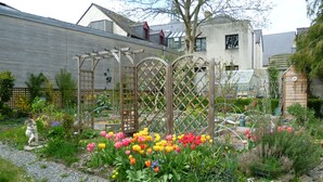 Un jardín con diversas flores y plantas, una pérgola de madera y una estatua.