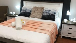 A neatly made bed with a white headboard, a fluffy pillow, and a peach-colored blanket. There is a bedside table with a lamp and a small decorative item. - Egrets Rest - Daintree (Daintree)