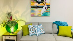 A living room with a sofa, decorative pillows, a wall-mounted painting of a bird, and a green glass lamp on a side table. - Egrets Rest - Daintree (Daintree)