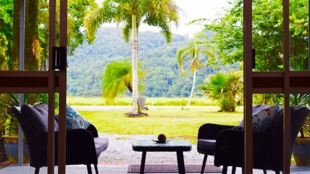 A room with a large window offering a view of a lush garden and a mountain. There are two chairs and a small table in the room. - Egrets Rest - Daintree (Daintree)