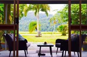 A room with a large window offering a view of a lush garden and a mountain. There are two chairs and a small table in the room. - Egrets Rest - Daintree (Daintree)