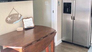 A kitchen with a stainless steel refrigerator, a wooden dining table with two stools, and a wall-mounted flat-screen TV. - SURF SHACK (Cape Canaveral)