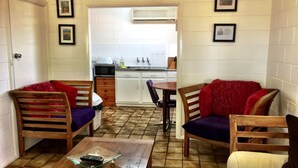 A compact living space with a kitchenette, two wooden chairs with red cushions, a small table, and a glass coffee table. - The Picker's Hut - Peaceful vineyard setting (Monash)