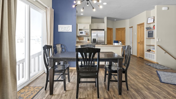 Un coin repas avec une table et des chaises en bois, un lustre moderne et, en arrière-plan, une vue sur la cuisine.