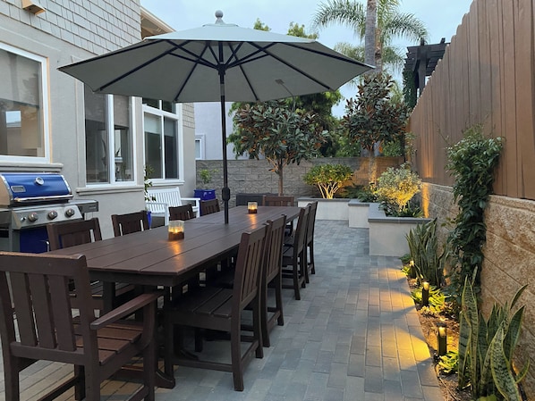 An outdoor patio with a dining table, chairs, a grill, and a large umbrella.