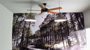 Een eetgedeelte met een muurschildering van een bos, een plafondventilator, hanglampen en een tafel gedekt met borden en een fruitschaal.