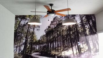 A dining area with a mural of a forest, a ceiling fan, pendant lights, and a table set with plates and a bowl of fruit.