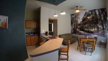 A modern kitchen with a central island, wooden dining table, and chairs. A large mural of a forest scene is on the wall.