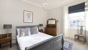 Une chambre à coucher avec un grand lit, une commode, une chaise, une table de chevet, une lampe, un miroir, un lustre et une fenêtre avec des rideaux.