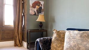 Une pièce avec un canapé bleu, des coussins à motifs, une lampe sur une table d’appoint en bois, un tableau au mur et une fenêtre avec des rideaux.