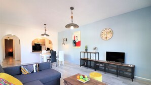 A living room with a sofa, a coffee table with fruit, a TV, and a dining area in the background.