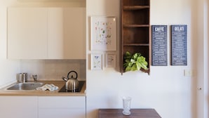 A kitchen with a sink, kettle, and a wall-mounted shelf with a plant. There are framed botanical prints and two signs listing coffee and gelato options. - Lago Terrione Apartment - Charme Holidays (Rome)