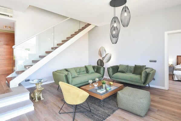A modern living room with a staircase, green sofas, a glass coffee table, and a yellow chair.