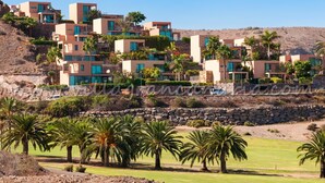 A modern resort with multi-story buildings, palm trees, and a rocky hillside backdrop. -  Los Lagos 39 Salobre Golf (Maspalomas)