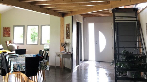 A spacious room with a high ceiling, wooden beams, a dining area with chairs and a table, and a staircase with a railing.