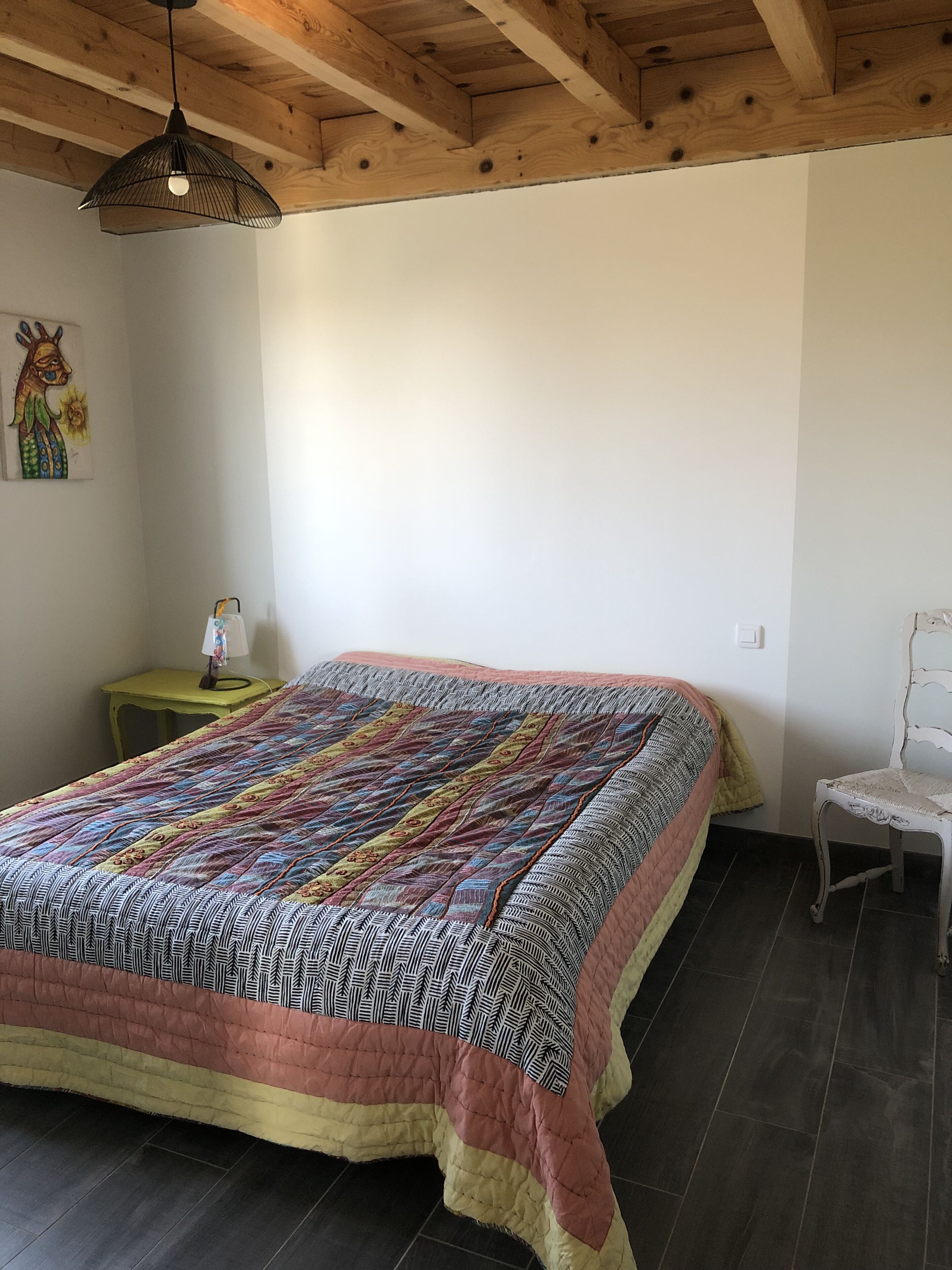 Een slaapkamer met een groot bed, een klein nachtkastje, een stoel en een houten plafond.