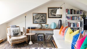 Un salon chaleureux avec un plafond haut, une bibliothèque remplie de livres, un canapé blanc agrémenté de coussins colorés, un fauteuil ancien et un tapis à motifs.
