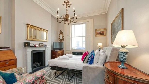 A hotel room with a bed, a sofa, a dresser, a television, a fireplace, and a chandelier. - Traditional 1 Bed Apartment, Pimlico (London)