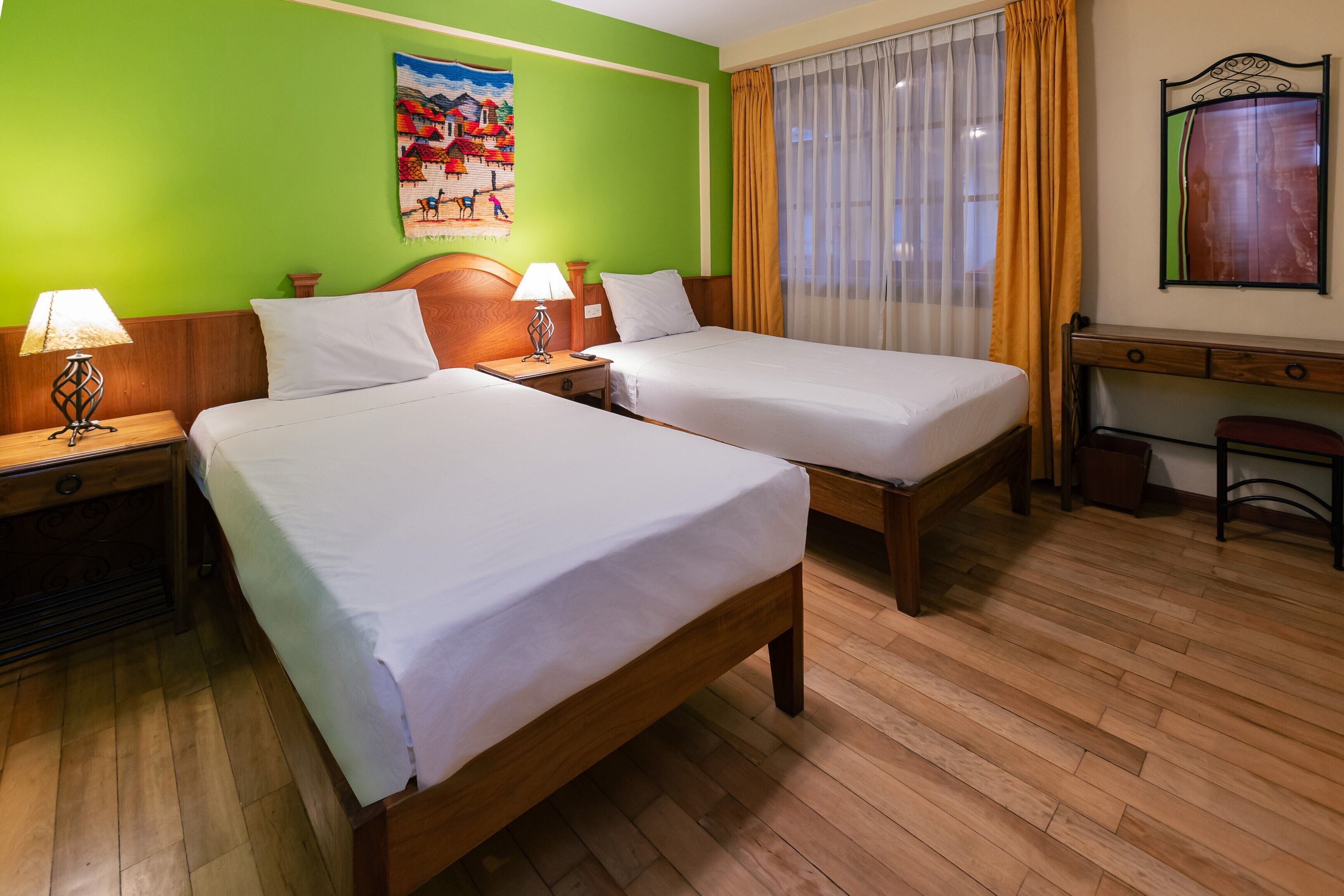 A hotel room with two single beds, wooden headboards, a desk with a chair, a small table, and a window with curtains.