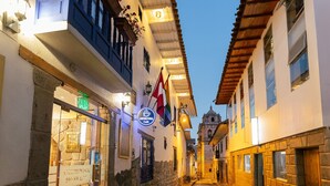A cobblestone street with buildings on either side, a café sign, and a church tower in the distance. - Amerindia Hotel (Cusco)