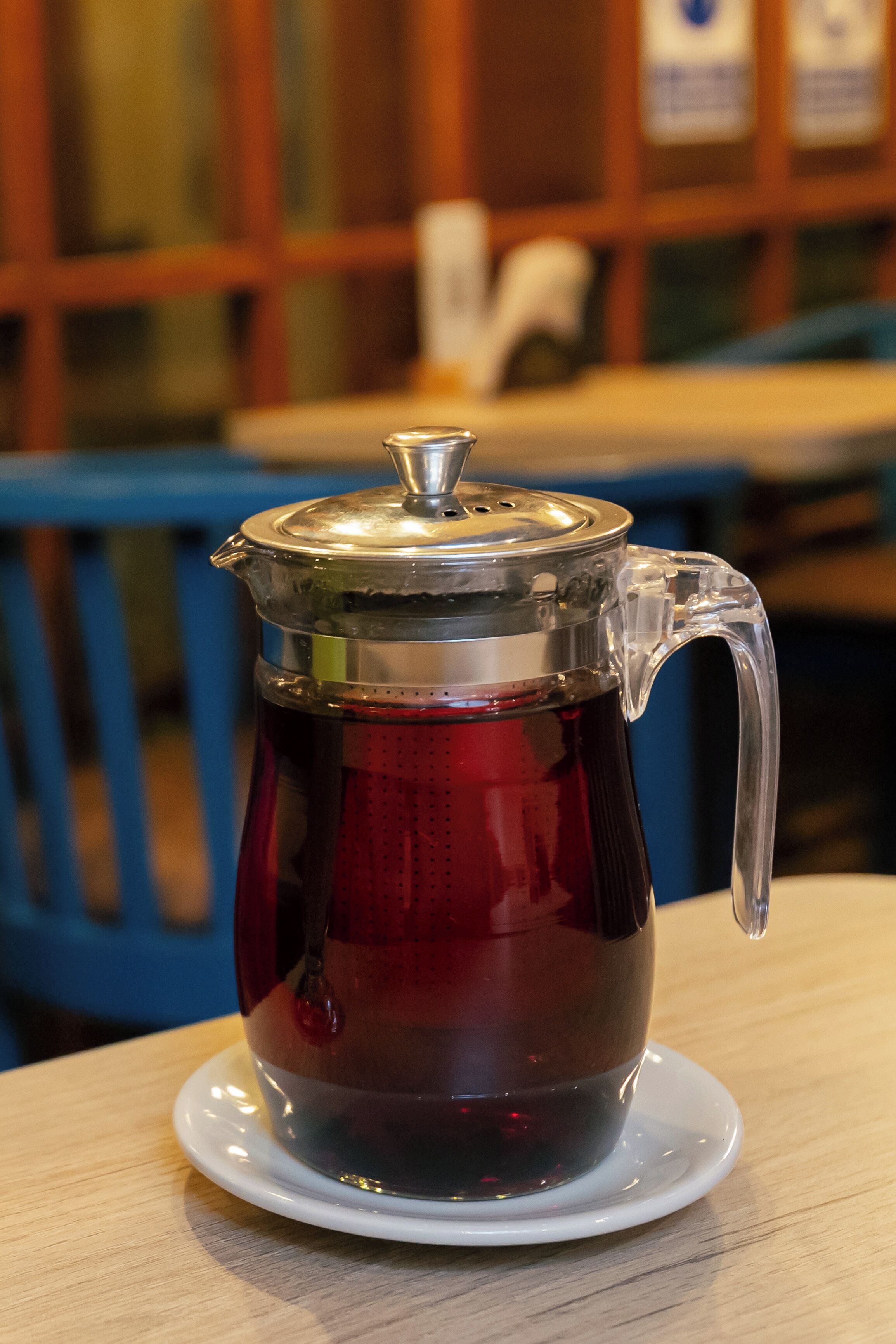 A clear glass teapot with a metal lid on a white saucer, placed on a wooden table.