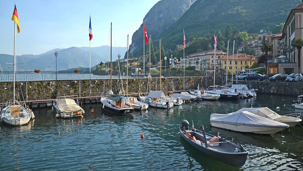 A marina with boats docked, flags, and a mountainous backdrop.