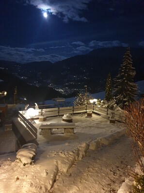 Zona exterior cubierta de nieve con bancos y una cerca, iluminada por luces ambientales.
