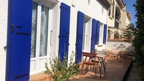 Un bâtiment aux murs blancs et aux volets bleus, avec une plante en pot et des sièges à l’extérieur.