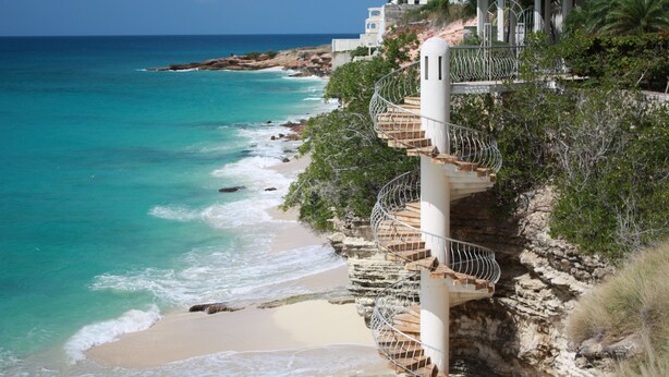 Ein Hotel an der Küste mit einer Wendeltreppe, die zu einem Balkon mit Blick auf türkisfarbenes Wasser und einen Sandstrand führt.