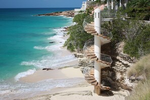 A coastal hotel with a spiral staircase leading to a balcony overlooking turquoise waters and a sandy beach.