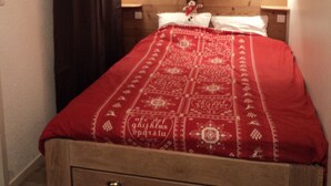 Quarto com cabeceira de madeira, duas luminárias de apoio, colcha vermelha e mesa de cabeceira pequena com abajur.