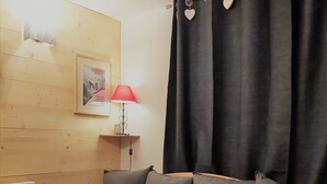 Quarto aconchegante com piso de madeira, um sofá com almofadas cinzas, luminária vermelha, um quadro na parede e cortina preta.