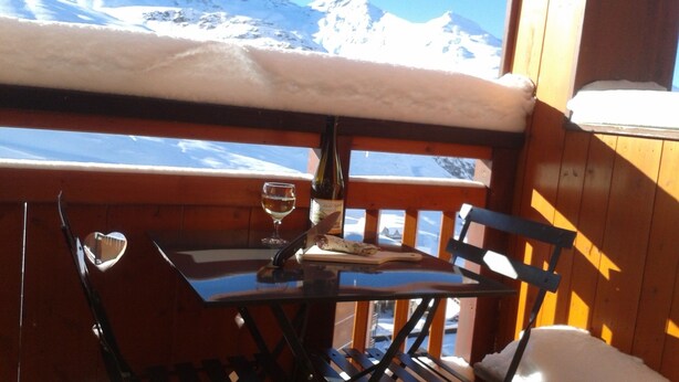 Uma mesa de madeira com uma taça de vinho e uma garrafa, uma cadeira e vista para montanhas cobertas de neve.