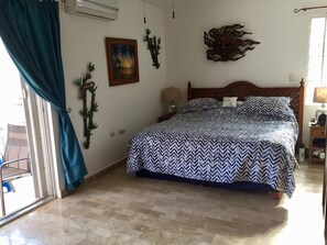 A bedroom with a bed, a window with curtains, a wall-mounted air conditioner, a wall decoration, and a nightstand with a lamp.