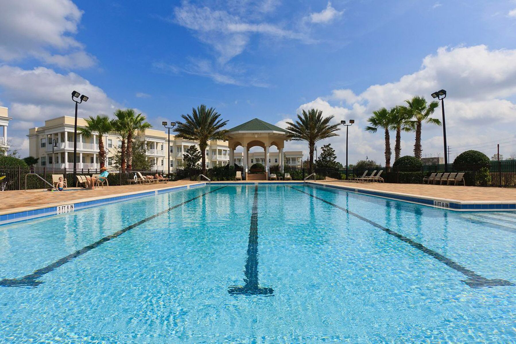 A clear swimming pool with lounge chairs and palm trees, surrounded by a building with a covered area.